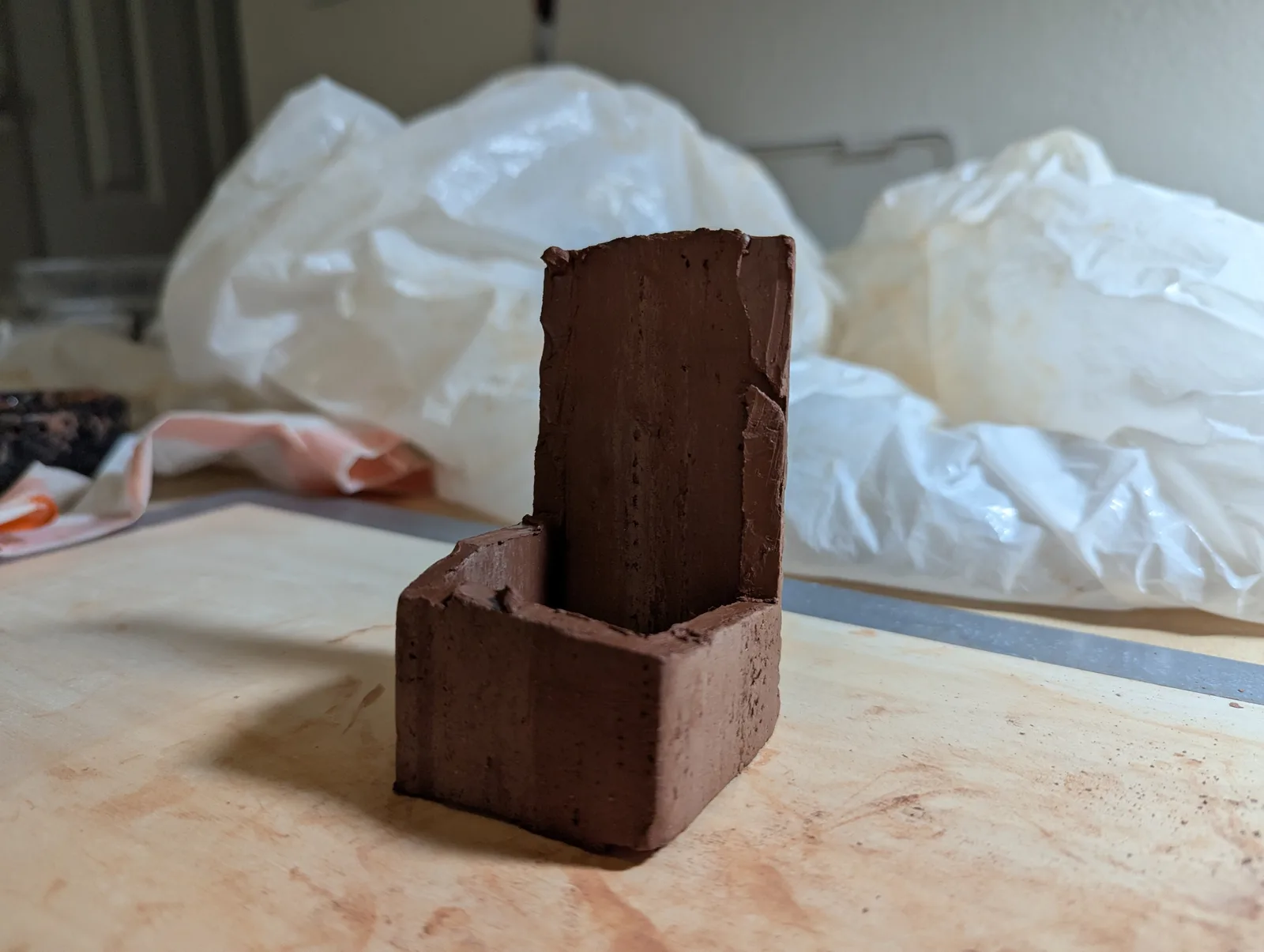 A clay sculpture in progress sits on a wooden surface with plastic sheeting in the background.