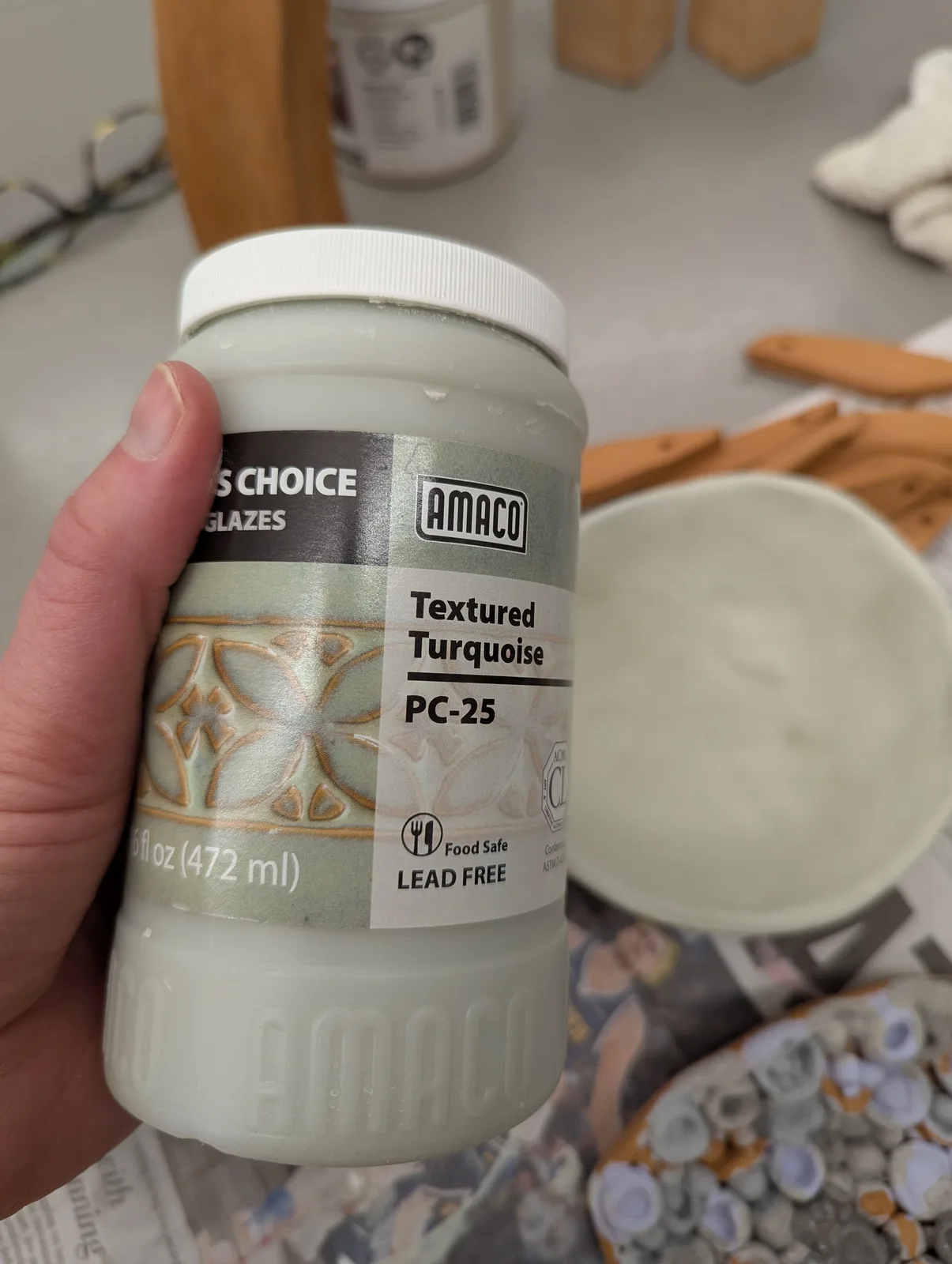 A hand holds a jar of Amaco Textured Turquoise glaze, with pottery tools and a bowl in the background.