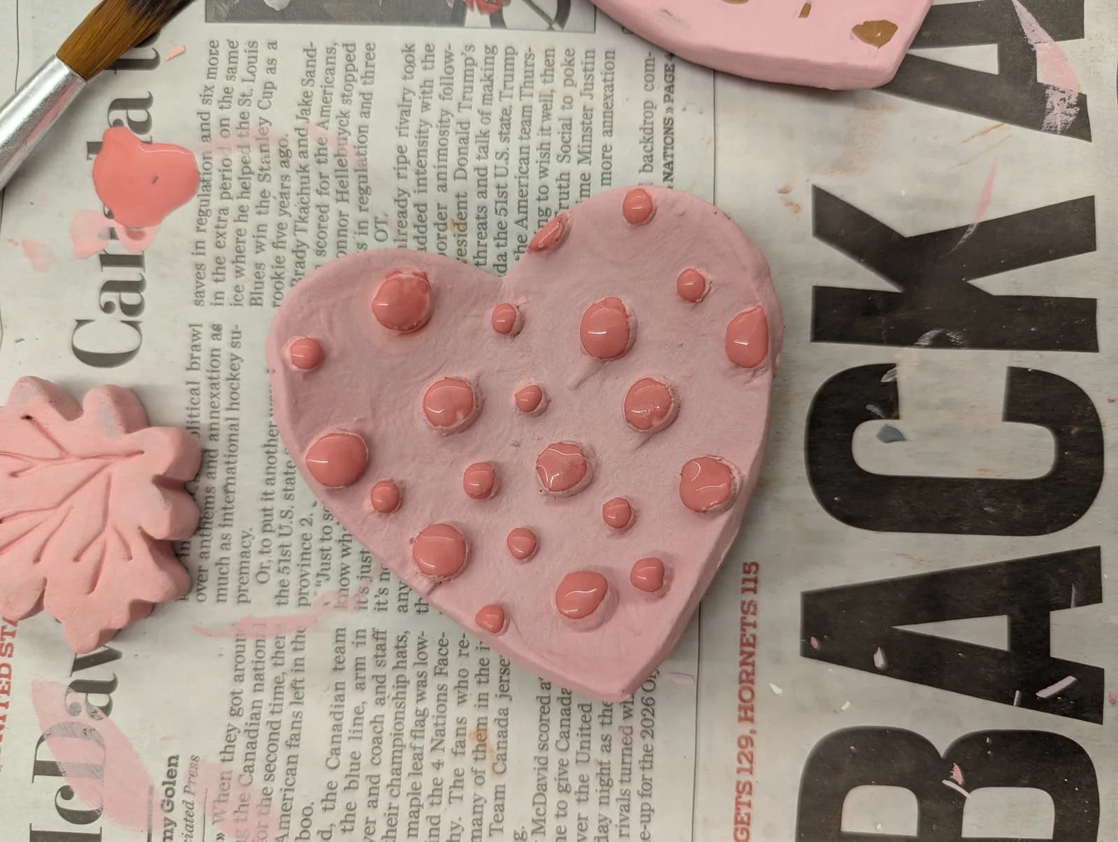 A pink clay heart with raised dots is shown in progress on newspaper with a paintbrush.