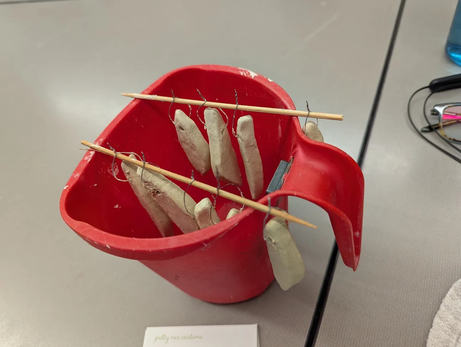 Clay pieces drying in a red container, suspended by wires and wooden sticks.