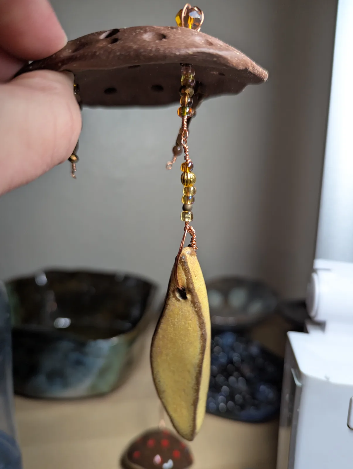 A hand holds a ceramic piece with beaded strands hanging from it, showing process.