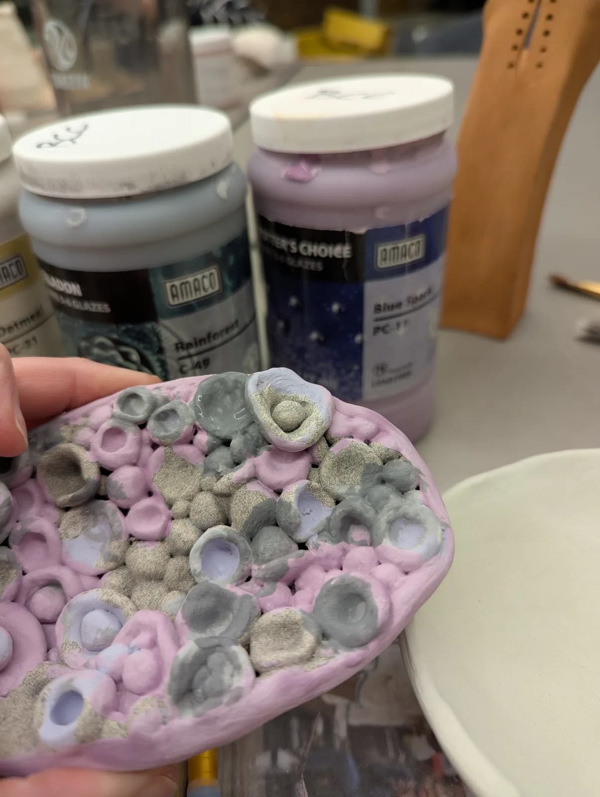A ceramic piece in progress with purple and gray glazes, surrounded by art supplies in a studio setting.