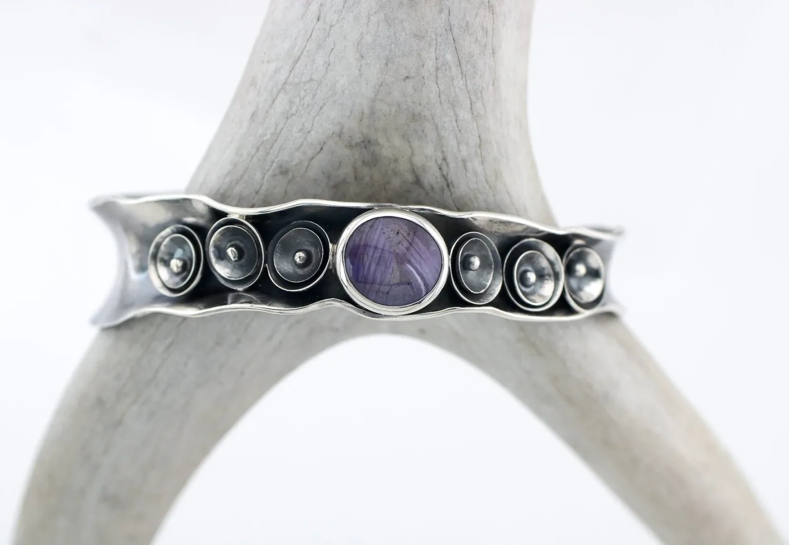 A silver cuff bracelet features a central purple stone surrounded by smaller circular metal elements.