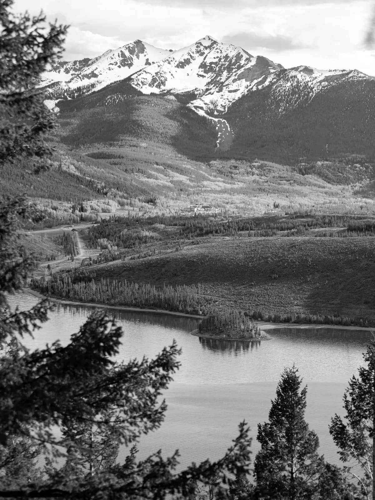 A black and white photograph captures a vast landscape with snow-capped mountains overlooking a serene lake and forested hills.