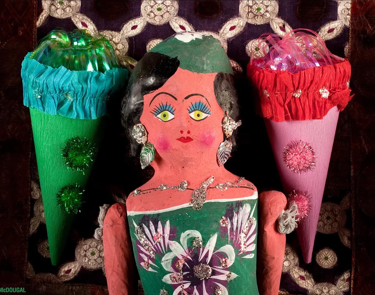 A folk art figure with decorative cones and jewelry, presented against a patterned textile.