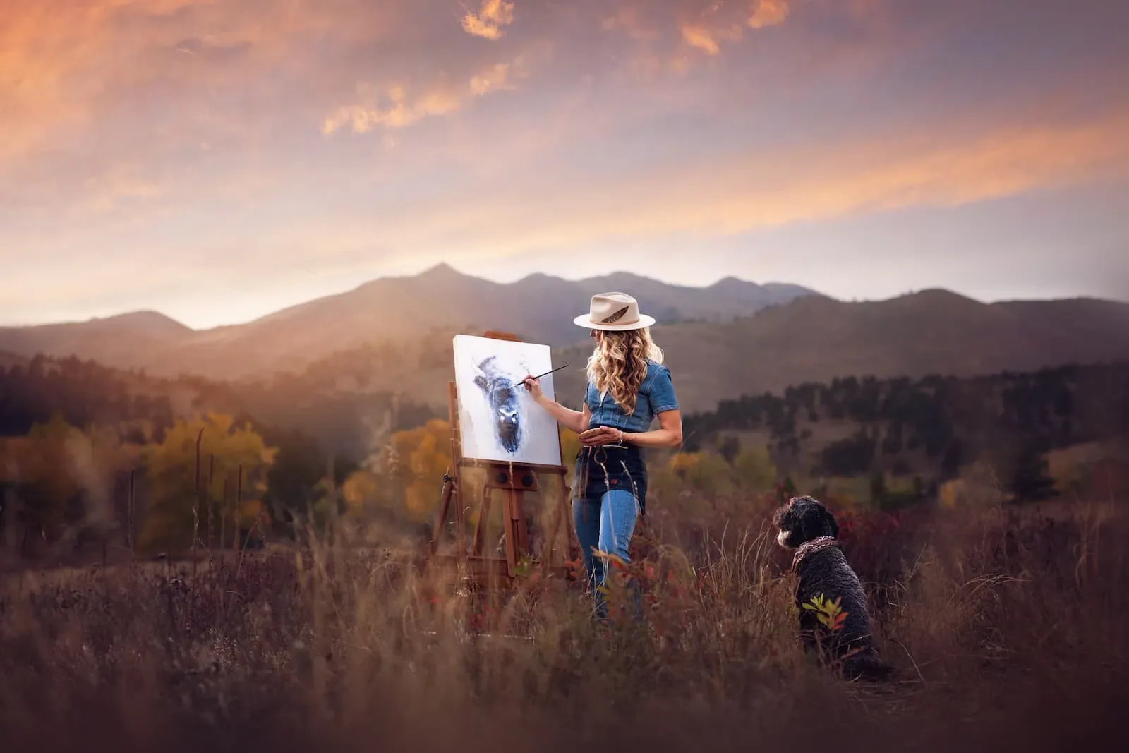 An artist paints a buffalo in a field with mountains and a dog nearby at sunset.