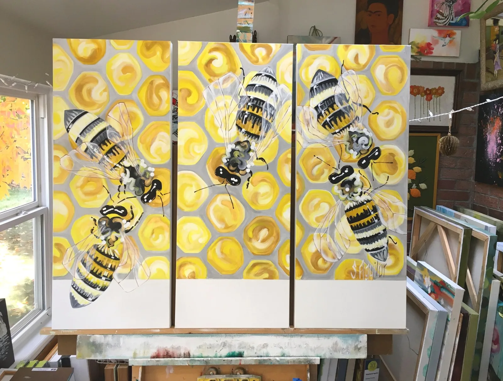 A triptych painting of bees on a honeycomb background is displayed in an artist's studio.