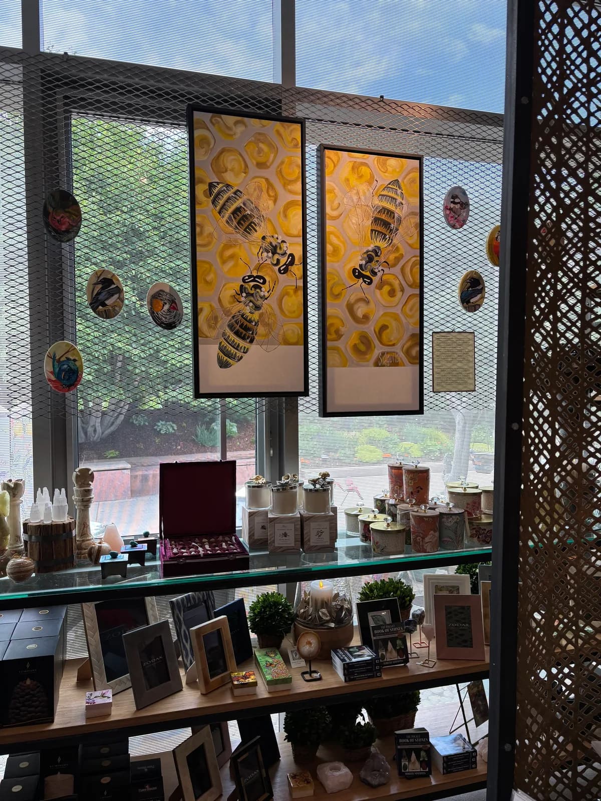 Two framed paintings of bees on a honeycomb background are displayed in a shop setting with various retail items.