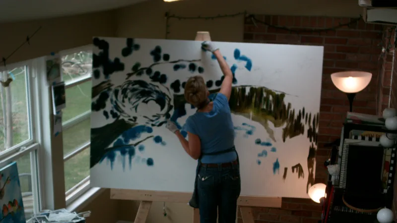 An artist works on a large abstract painting in a studio with a window and brick wall.