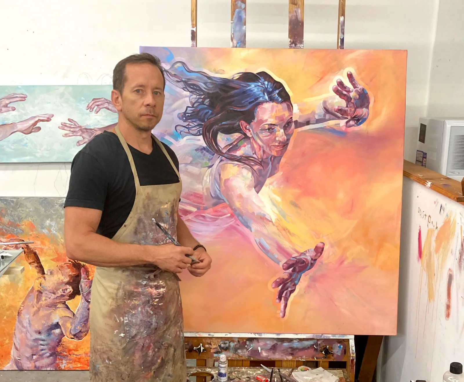 An artist stands in his studio next to a vibrant, expressive painting of a figure in motion.