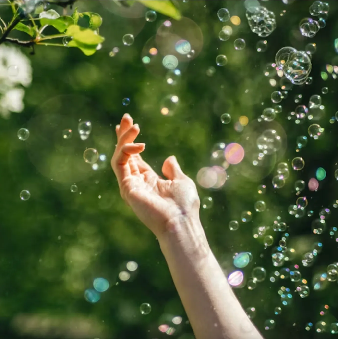 A hand reaches out towards a cascade of iridescent bubbles against a lush green, natural background.