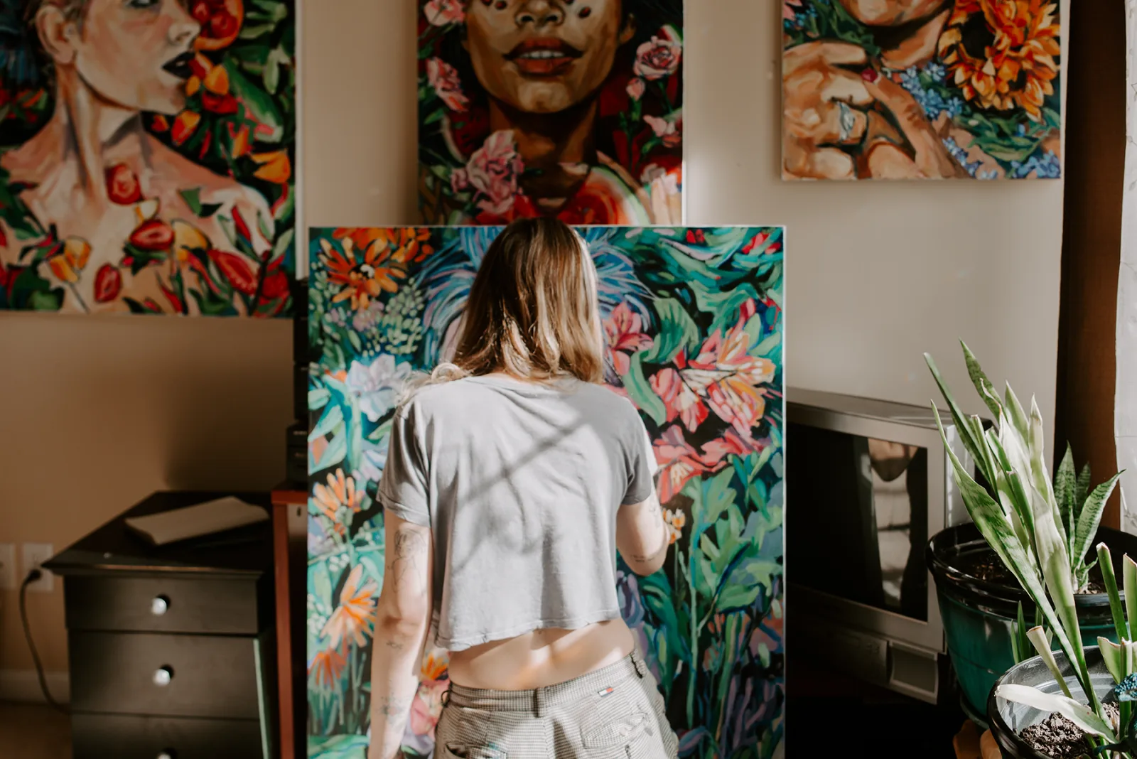 An artist works on a vibrant floral painting in a studio setting with other artworks visible.