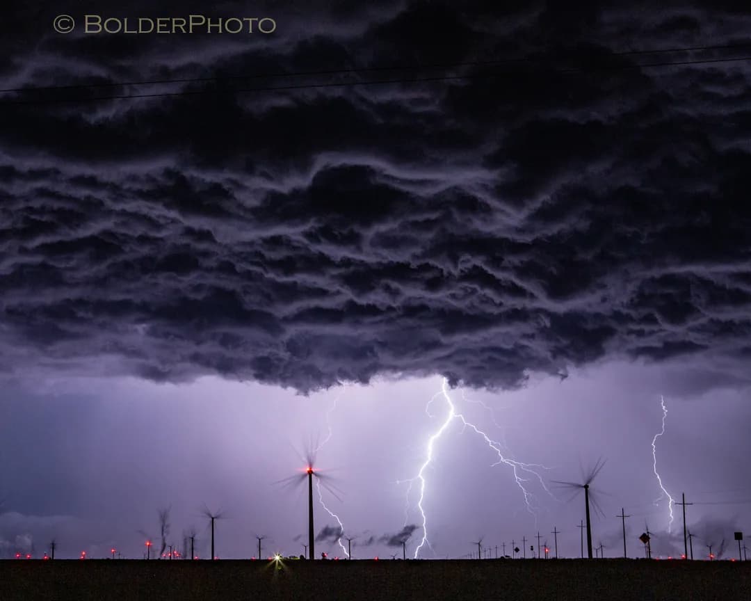A dramatic photograph captures lightning striking behind wind turbines under a dark, turbulent sky.