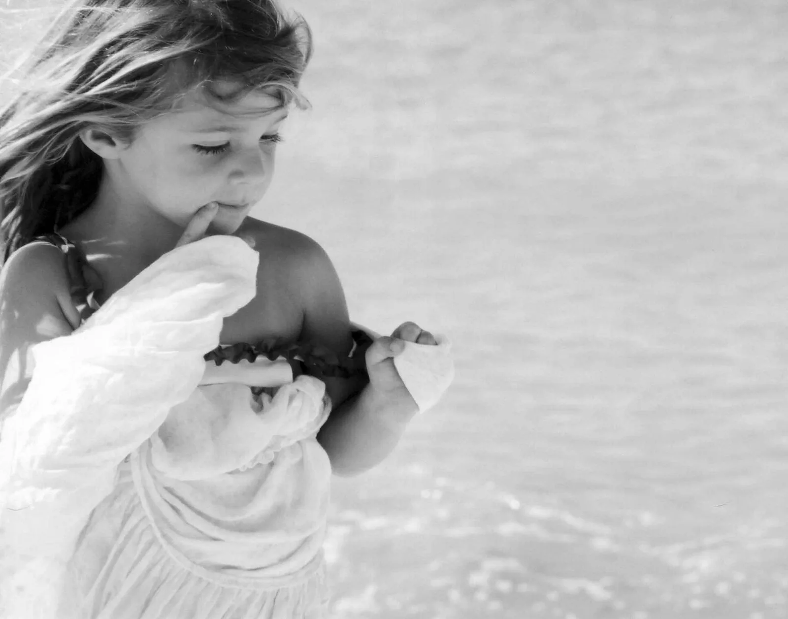 A black and white photograph captures a young girl with her finger to her lips, holding a piece of fabric near the water.