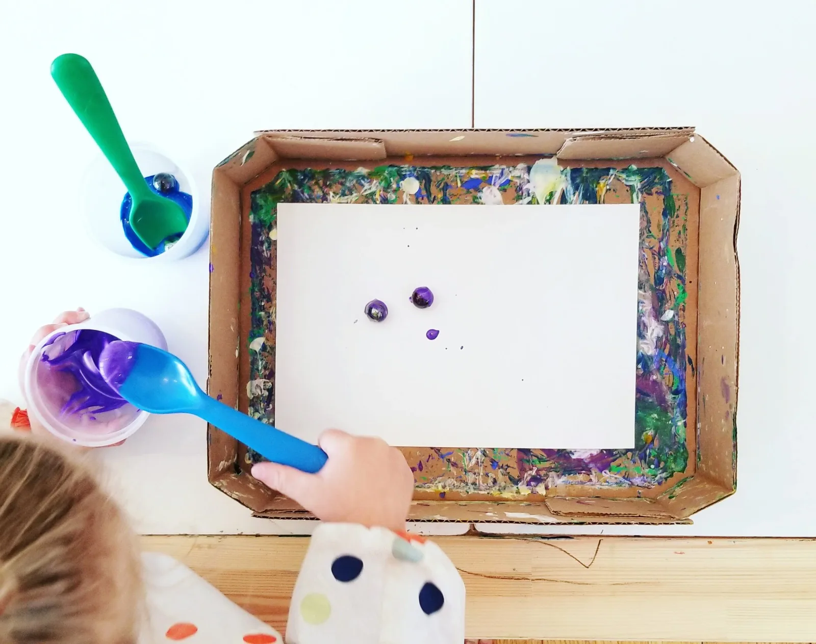 A child's hands add purple paint to a marbling art project with marbles and spoons.
