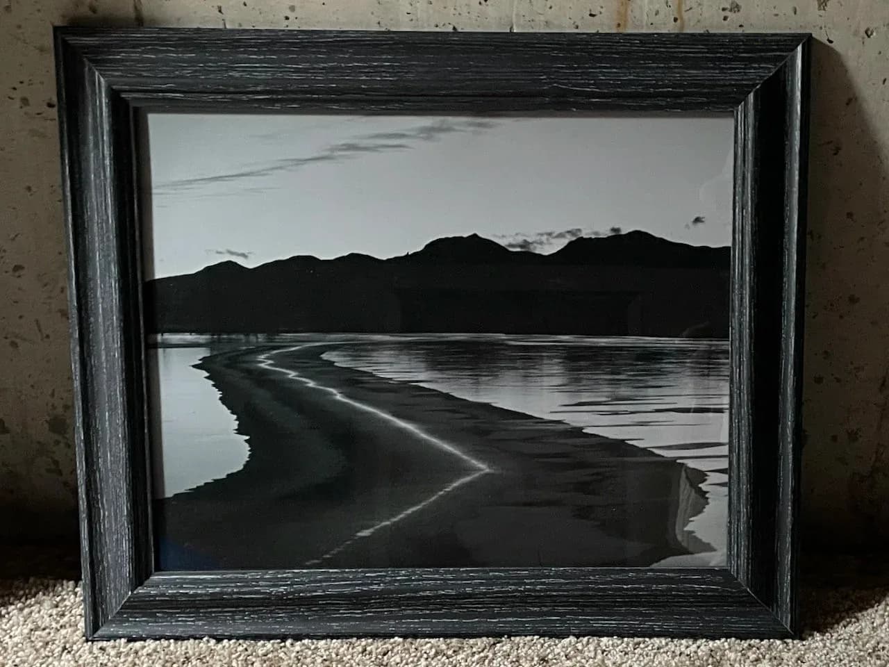 A black and white photograph captures a serene landscape with mountains reflected in calm water.