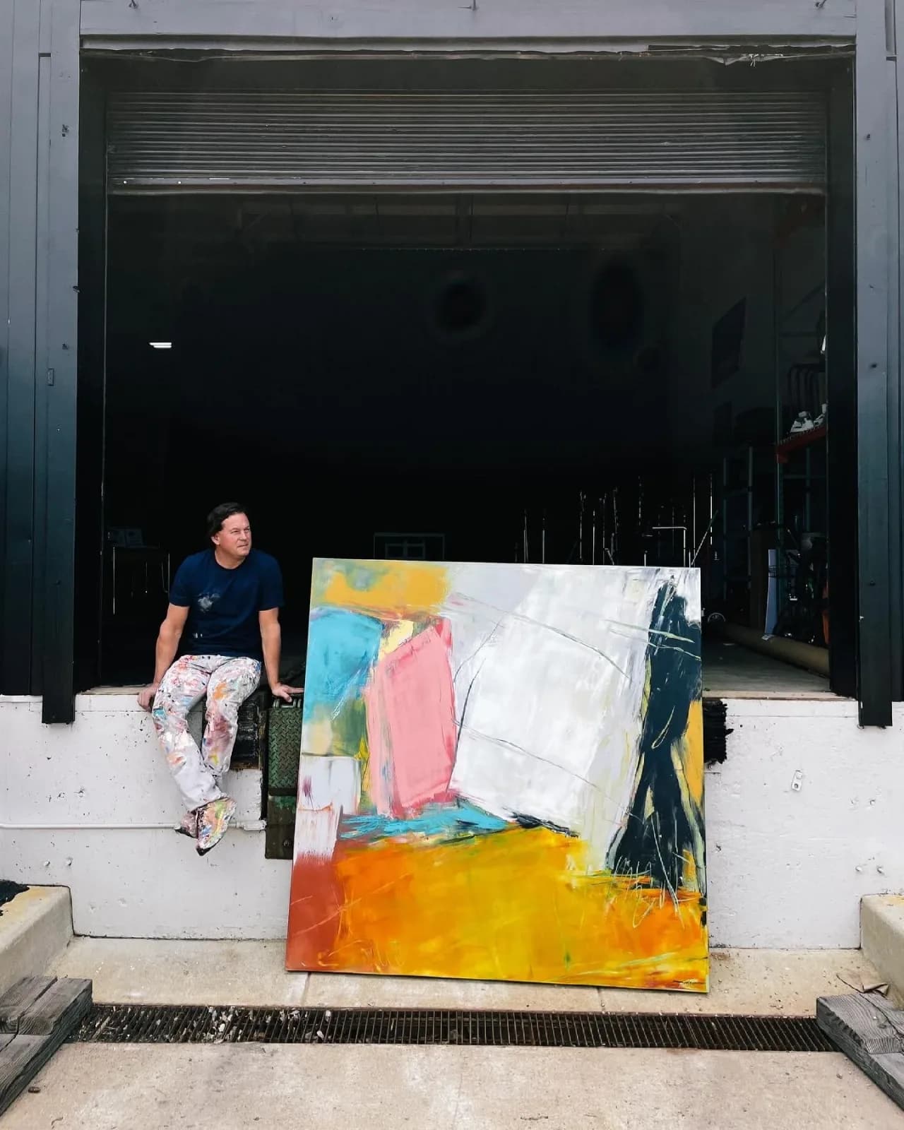 An artist sits beside a large abstract painting in an industrial setting.