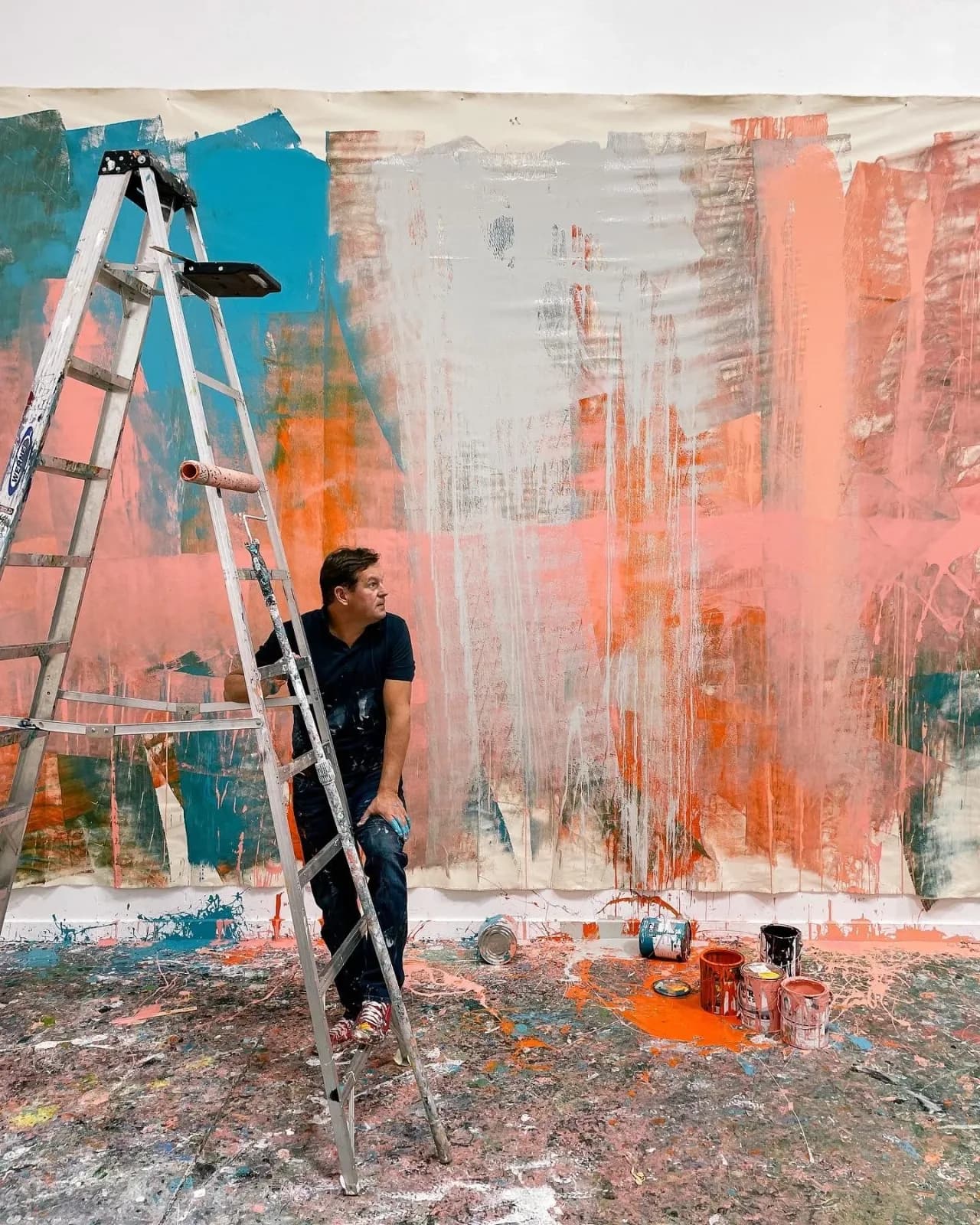 An artist stands on a ladder in a studio with a large abstract painting and scattered paint cans.