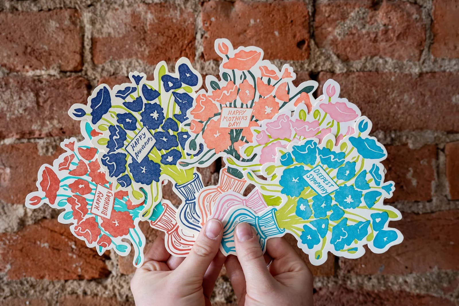 A set of folk-art floral greeting cards in shades of East Bay, Pelorous, and Cloud are presented against a brick background.