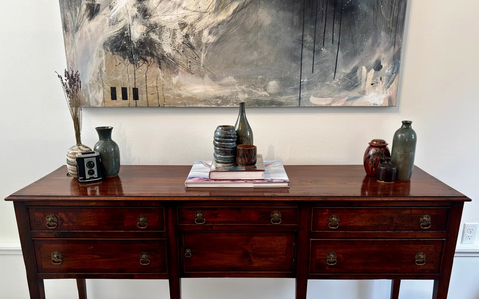 An abstract painting in shades of gray and tan hangs above a wooden sideboard displaying decorative pottery and a vintage camera.