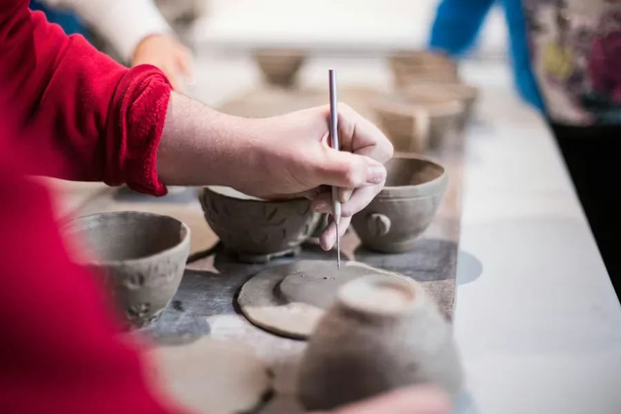 A person's hands work on unfinished clay pottery, including bowls and cups, on a table in a workshop.