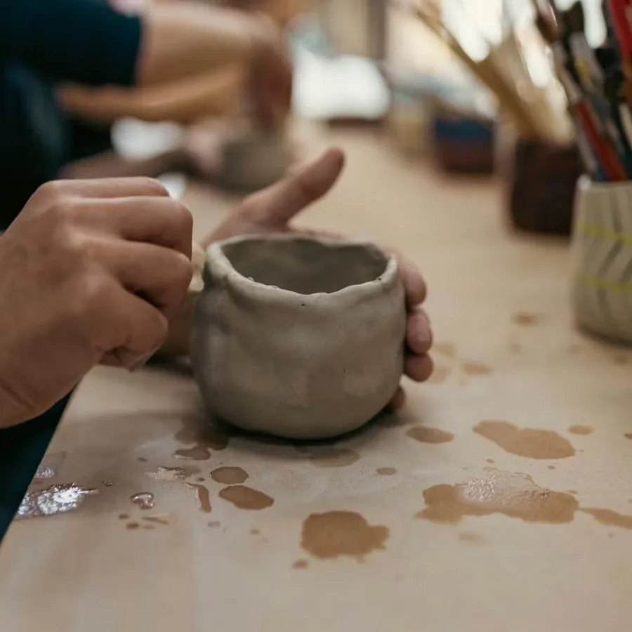 Hands shaping a clay pot on a table in a pottery studio with brushes in the background.