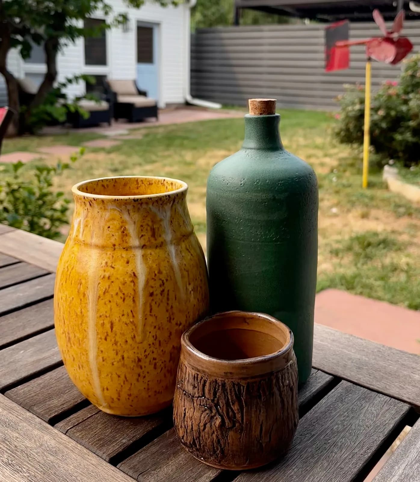 Three ceramic vessels, including a yellow speckled vase and a green bottle, are displayed on a wooden table in a garden setting.