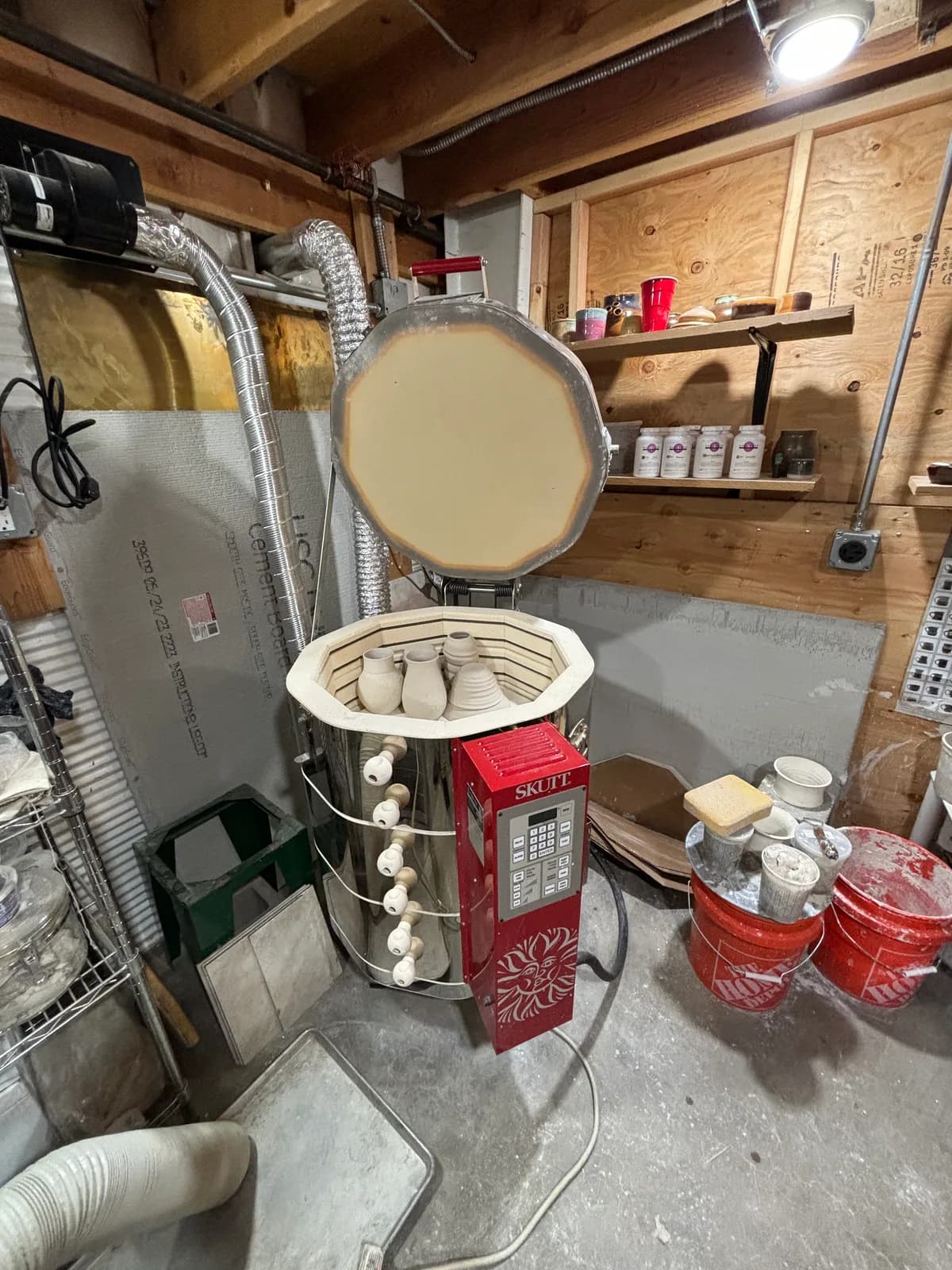 A pottery kiln filled with small ceramic vessels sits in a furnished studio with shelves and buckets.