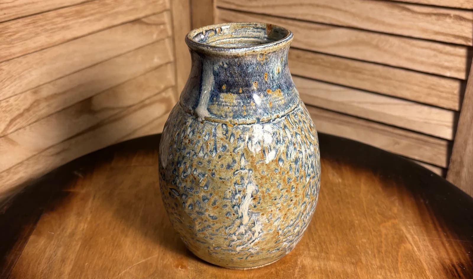 A ceramic vase with a speckled glaze in earthy tones and blue accents rests on a wooden surface.