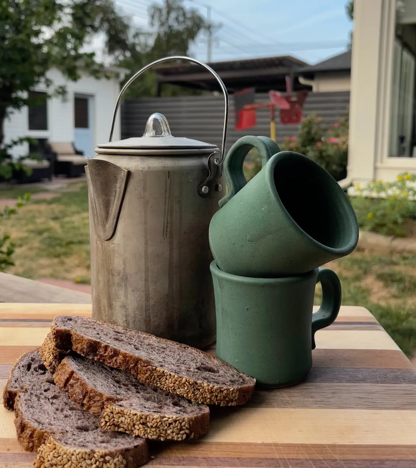 A ceramic kettle and two mugs are displayed with sliced bread on a wooden cutting board in a backyard setting.