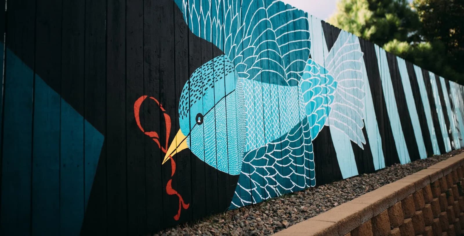 A vibrant mural of a blue bird with intricate patterns is painted on a black wooden fence.